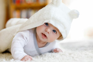 Baby girl with blue eyes wearing white towel or winter overal in white sunny bedroom. Newborn child relaxing in bed. Nursery for children. Textile and bedding for kids. New born kid with toy bear.